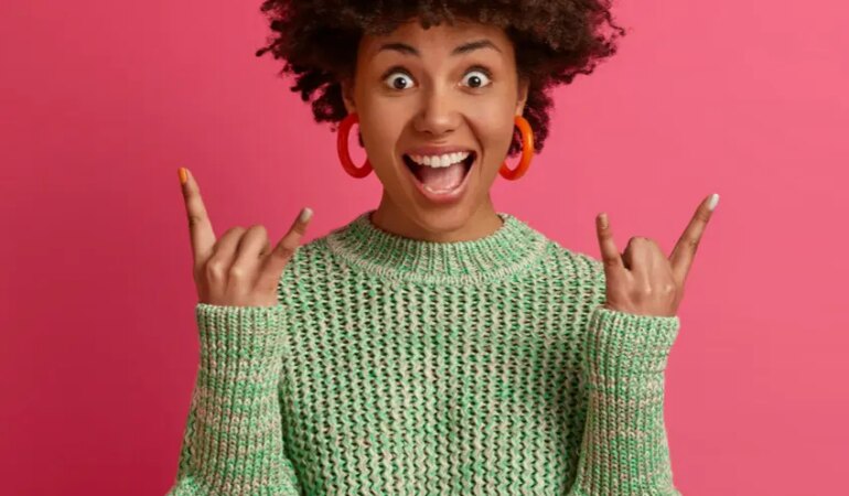 happy carefree dark skinned rebellious young woman enjoys awesome music makes rock n roll gesture has fun on music festival or cool event wears casual jumper poses against pink wall